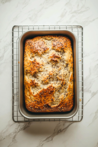 This image shows the baked bread loaf resting in the pan on a wire rack placed over a white marble countertop.