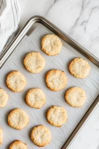 baked-shoThis image shows a top-down view of a silver baking sheet on a white marble countertop, with freshly baked shortbread cookies featuring lightly golden edges and no background clutter.rtbread-cookies-on-baking-sheet-with-golden-edges-on-white-marble