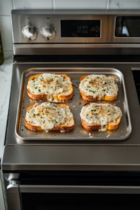 This image shows four slices of French bread topped with cheese and garlic butter inside an oven, with cheese just starting to melt, placed on a white marble cooktop.