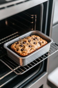 This image shows a chocolate chip pound cake loaf baking in a silver loaf pan placed on the center oven rack, captured through an open oven door from a top-down angle.
