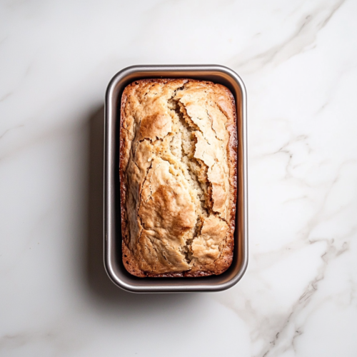 This image shows a golden-brown banana loaf cake in a shiny silver loaf pan on a clean white marble countertop, captured from a top-down angle.