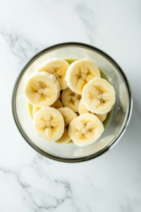 This image shows a tall clear glass on a white marble countertop with thin banana slices neatly stuck along the inside wall of the glass.