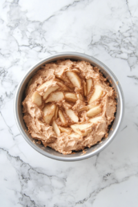 This image shows layers of cake batter and cinnamon-sugar apples inside a greased Bundt pan on a white marble countertop.