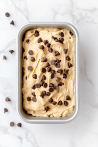 This image shows a top-down view of chocolate chip pound cake batter spread evenly in a greased silver loaf pan on a white marble countertop.