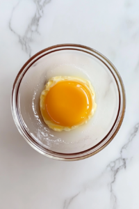 This image shows a small clear glass bowl with beaten eggs sitting on a white marble countertop.