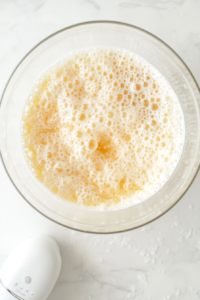 This image shows a glass mixing bowl containing beaten eggs, which are light yellow and frothy, placed on a white marble countertop.