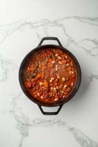 black-dutch-oven-with-diced-tomatoes-black-beans-pinto-beans-vegetable-broth-bay-leaf-on-white-marble-countertop