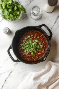 black-dutch-oven-with-homemade-chili-stirred-with-chopped-cilantro-vinegar-and-salt-on-white-marble-countertop