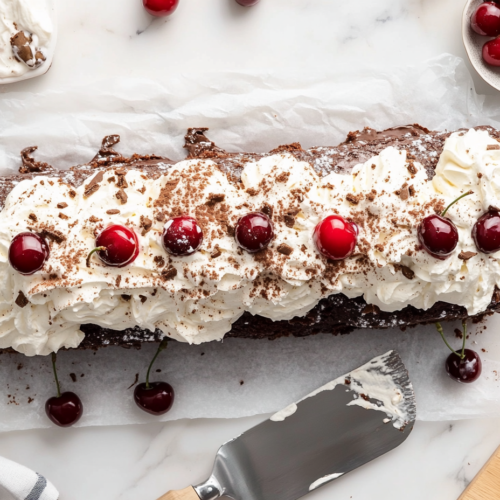This image shows a beautifully sliced Black Forest cake roll on a white serving platter, garnished with whipped cream peaks and cherries, over a clean white marble cooktop.
