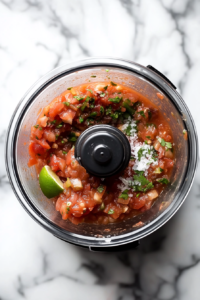 This image shows a clear food processor bowl filled with freshly blended salsa of a chunky texture. Ingredients like tomato, onion, jalapeño, and cilantro are visible within the blend.