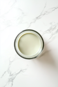 This image shows a clear glass blender jar on a white marble countertop, blending the avocado, banana, honey, and milk into a smooth light green mixture.