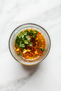 This image shows a small glass blender cup on a white marble countertop, filled with chili-lime dressing ingredients including lime juice, olive oil, minced garlic, chopped cilantro, honey, chili flakes, and salt before blending.