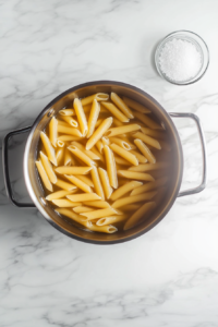 This image shows a large stainless steel pot filled with boiling salted water and fusilli pasta cooking, with a small glass bowl of reserved pasta water nearby, all on a clean white marble cooktop.