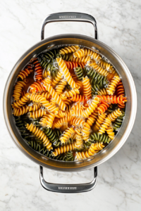 This image shows rotini pasta boiling in a large stainless steel pot filled with water, sitting on a white marble cooktop.