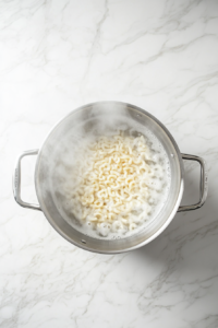 This image shows a top-down view of uncooked elbow macaroni added to boiling water inside a stainless steel pot on a clean white marble cooktop, with no background clutter.
