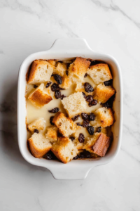 This image shows cubed bread arranged in a white dish, with melted butter and raisins added on top, ready for the pudding mixture. 📸 STEP 3 – Mix Custard Ingredients Image Name: whisked-custard-mixture-in-glass-bowl-over-white-marble-counter Prompt: Top-down view of a medium glass mixing bowl placed on a clean white marble cooktop, containing a well-whisked custard mixture made from milk, granulated sugar, 2 beaten eggs, ground cinnamon, and vanilla extract. The ingredients are fully blended, with no utensils or other items in the background. iPhone 16 Pro Max --ar 4:6 Alt Text: This image shows a smooth custard mixture in a glass bowl made from milk, sugar, eggs, cinnamon, and vanilla, ready to be poured over the bread. 📸 STEP 4 – Pour Custard and Soak Bread Image Name: custard-poured-over-bread-in-white-dish-on-white-marble Prompt: Top-down view of a white 8-inch square ceramic baking dish on a clean white marble cooktop. The custard mixture is already poured over the cubed bread and raisins, soaking in. A metal fork rests on the side, used for pressing the bread down to ensure it absorbs the custard. No extra ingredients or utensils shown. iPhone 16 Pro Max --ar 4:6 Alt Text: This image shows custard mixture poured over the bread and raisins in a baking dish, with a fork used to press it down for full soaking. 📸 STEP 5 – Baked and Ready to Serve Image Name: baked-bread-and-raisin-pudding-ready-to-serve-on-white-marble Prompt: Top-down view of the fully baked bread and raisin pudding in the white ceramic dish placed over a clean white marble cooktop. The surface is golden-brown with a crisp texture and visible raisins. No additional items or hands in the scene. Natural lighting makes the pudding look warm and inviting. iPhone 16 Pro Max --ar 4:6 Alt Text: This image shows the finished bread and raisin pudding after baking, with a golden crust and evenly distributed raisins, ready to be served. Let me know if you'd like me to adapt this format for other recipes too, Archie!