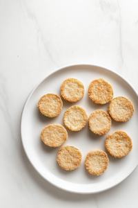 This image shows breaded zucchini slices arranged in a single layer on a white ceramic plate over a white marble countertop.