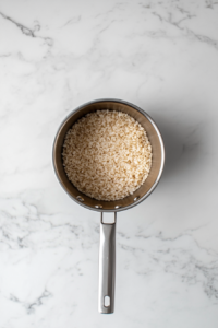 This image shows rinsed brown rice placed in a medium stainless steel saucepan over a white marble cooktop.