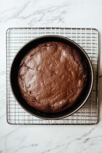 This image shows a chocolate cake resting on a wire cooling rack over a white marble cooktop.
