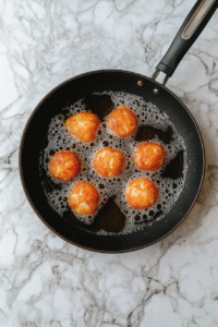 This image shows cheese bites frying to a golden color inside a black skillet placed on a white marble cooktop.