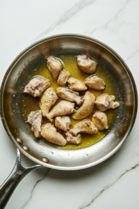 This image shows seasoned chicken pieces cooking in olive oil in a large stainless steel skillet on a clean white marble cooktop, with some pieces golden brown and others still cooking.