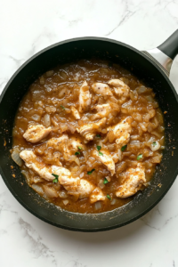 chicThis image shows raw chicken pieces added to a spiced onion mixture in a black skillet over a white marble cooktop.ken-pieces-added-to-spiced-onion-mixture-in-skillet