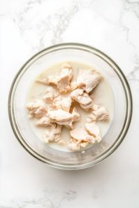 This image shows raw chicken pieces sitting inside a large clear glass bowl filled with white buttermilk, set on a clean white marble countertop, with no other items or ingredients in the background.