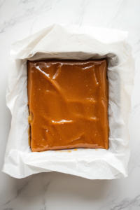 This image shows hot caramel being poured into a parchment-lined baking dish on a white marble cooktop, ready to cool.