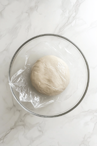 This image shows a top-down view of a glass bowl with pale cookie dough, covered in plastic wrap, chilling on a white marble countertop.