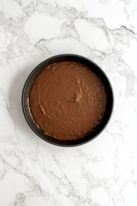 This image shows a round non-stick cake pan over a white marble cooktop filled with chocolate cake batter, ready to bake.