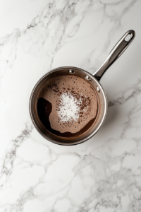 This image shows a smooth, velvety mixture of melted dark chocolate with milk, cream, powdered sugar, and espresso powder in a stainless steel saucepan on a white marble cooktop.