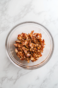 This image shows a glass bowl filled with chopped crispy bacon pieces, placed on a clean white marble cooktop.