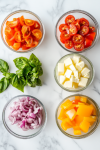 This image shows chopped red and yellow bell peppers, halved cherry tomatoes, diced red onions, pepperoni slices, mozzarella cubes, and fresh basil leaves in small clear glass bowls on a white marble surface.