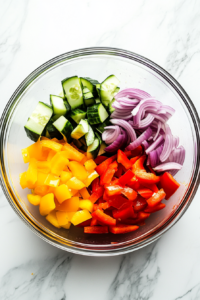 This image shows a top-down view of a clear glass bowl on a white marble countertop, containing freshly chopped red onion, red bell pepper, yellow bell pepper, and cucumber, ready for a dense bean salad.