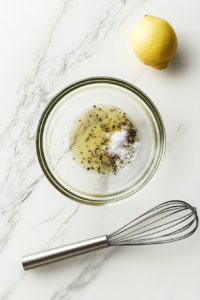 This image shows marinade ingredients like olive oil, lemon juice, garlic, oregano, salt, and pepper combined in a glass bowl on a white marble countertop.