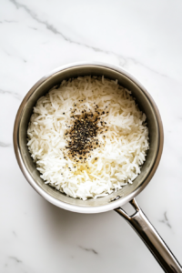 This image shows rice, salt, oregano, pepper, and water combined in a stainless steel saucepan on a white marble countertop.