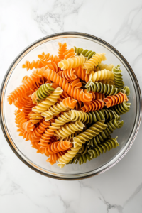 This image shows cooked and cooled rotini pasta sitting in a large clear glass bowl on a clean white marble cooktop.