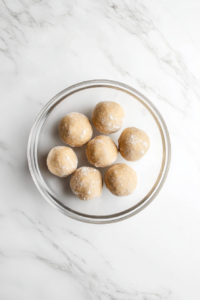 This image shows a top-down view of cookie dough balls rolled in sugar inside a glass bowl on a white marble countertop.