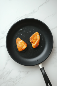 This image shows two chicken breasts frying in avocado oil in a black skillet on a white marble cooktop.