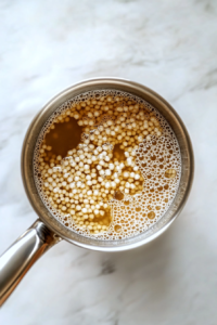 This image shows a top-down view of a stainless steel saucepan on a white marble countertop, with couscous cooking in boiling water and steam rising from the pot.