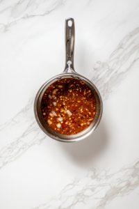 This image shows a small stainless steel saucepan on white marble cooktop simmering homemade ranchero sauce made with tomatoes, onions, garlic, and chili.