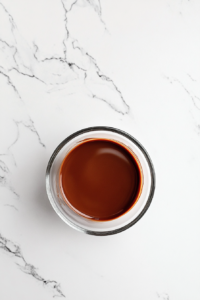 This image shows a glass bowl with a glossy melted mixture of chocolate and butter, set aside to cool on a white marble cooktop.
