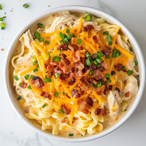 This image shows a large white ceramic bowl filled with creamy crack chicken noodle soup, topped with shredded cheddar cheese, crumbled bacon, and chopped green onions, placed on a clean white marble countertop with no other items in the background.