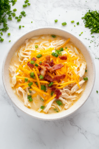 This image shows a serving of crack chicken noodle soup in a white ceramic bowl on a white marble countertop, topped with shredded cheddar, crumbled bacon, and chopped green onions.