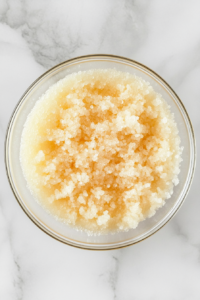 This image shows a clear glass bowl on a white marble countertop with a light and fluffy mixture of softened butter and turbinado sugar.