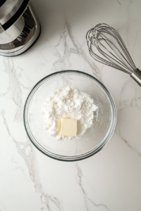 This image shows sugar and softened butter being creamed together in a glass bowl with an electric mixer, set on a white marble cooktop.