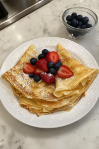 This image shows folded crepes served on a white plate with fresh strawberries, blueberries, and a dusting of confectioners’ sugar, with a glass bowl of batter on a white marble cooktop.