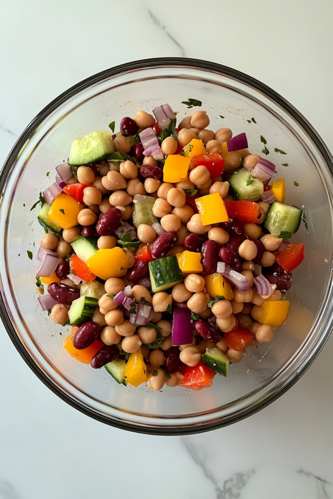 This image shows a top-down view of a clear glass bowl on a white marble countertop, containing freshly chopped red onion, red bell pepper, yellow bell pepper, and cucumber, ready for a dense bean salad.