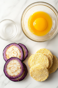 This image shows clear glass bowls with beaten egg and breadcrumbs, and sliced eggplant on a white marble countertop, ready for dipping and coating.