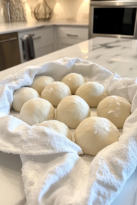 This image shows twelve dough balls covered with a white towel on a floured white marble countertop.
